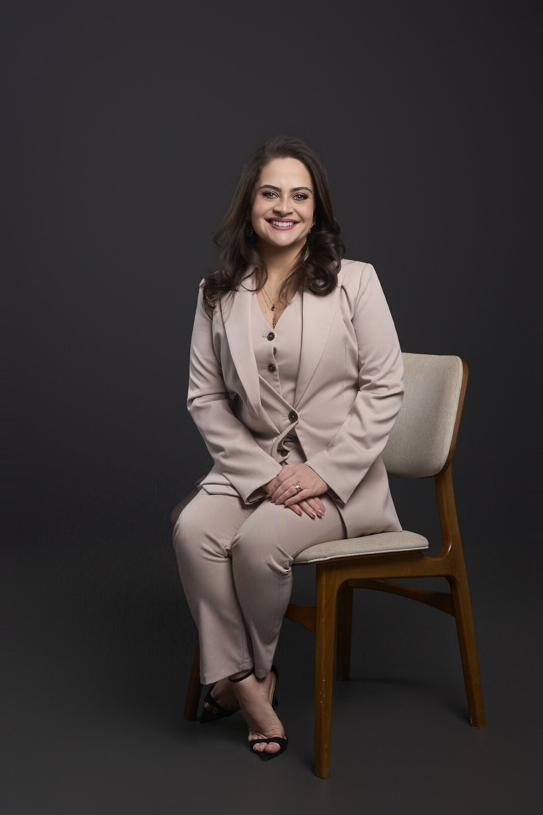 Dra. Claryssa Nascimento sorrindo, sentada em cadeira de madeira clara, com terno bege claro